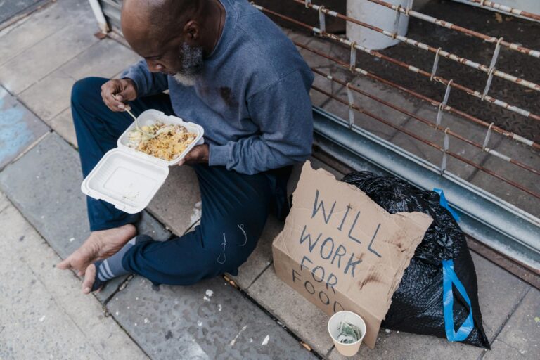 African American man sitting on a sidewalk, eating with a "Will Work for Food" sign.